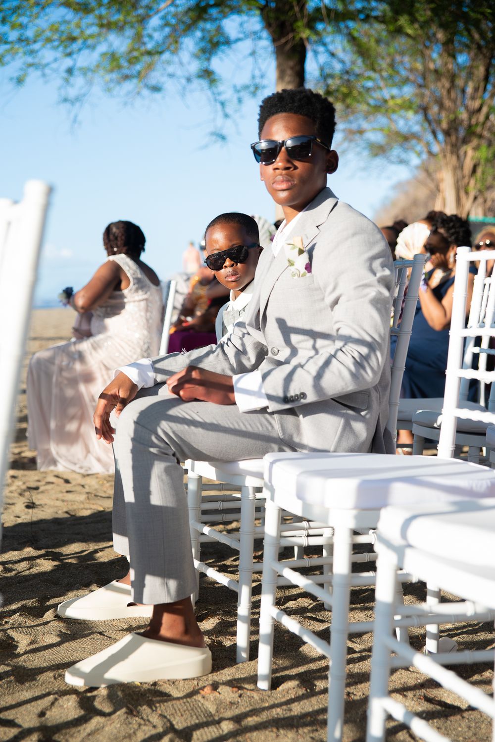 Young wedding guest in white suit and sunglasses sits in beach chair at outdoor ceremony.