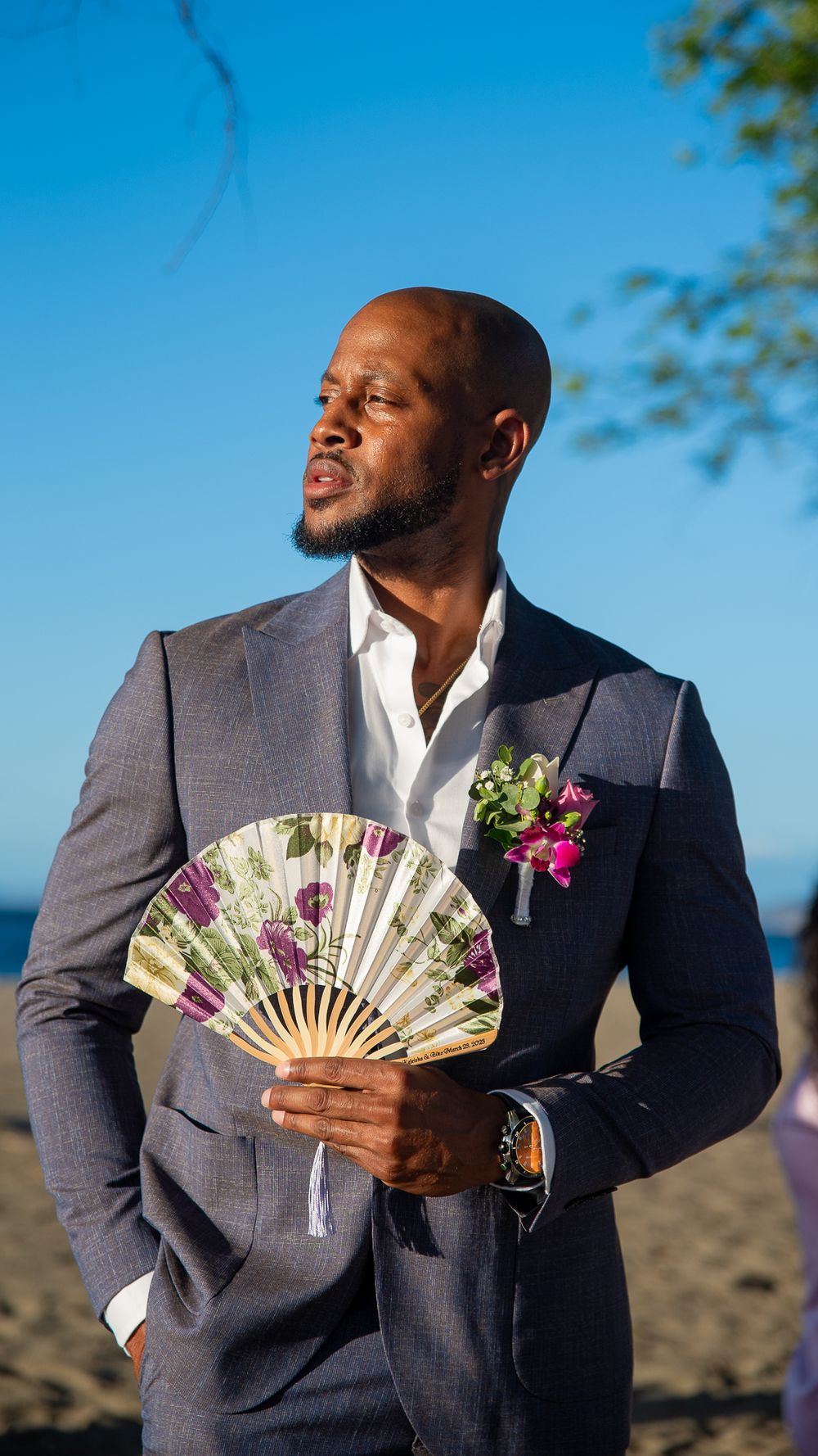 A person in a navy suit poses with a decorative fan against a bright blue sky backdrop.