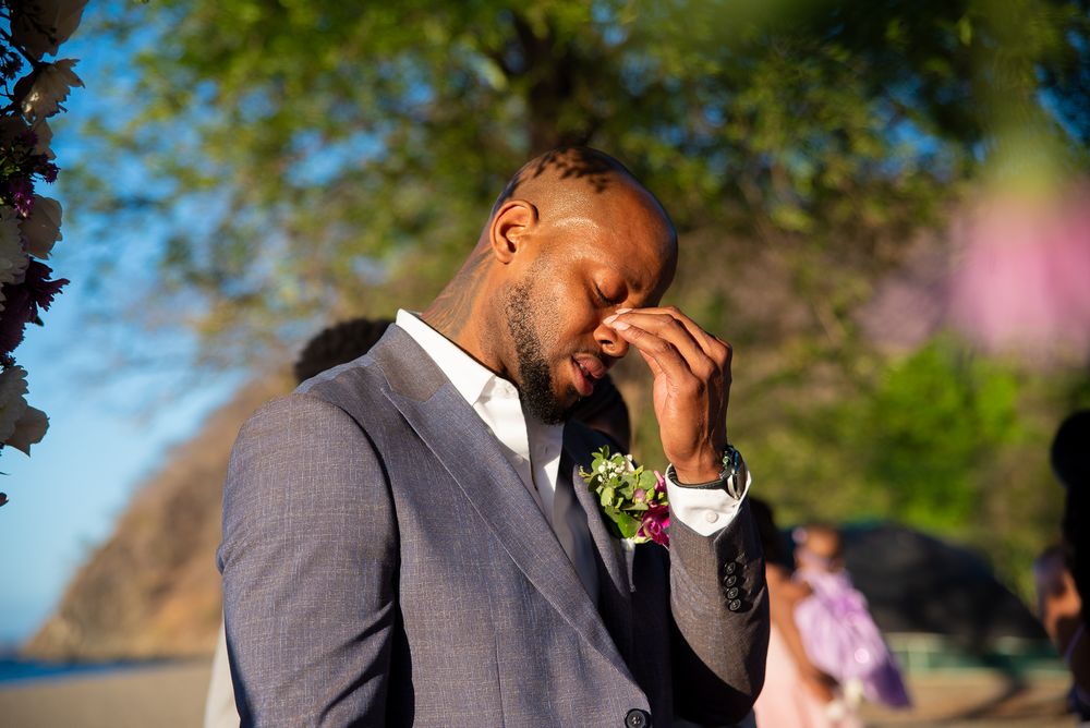 A person in a grey suit laughs joyfully while holding a floral-decorated fan outdoors.