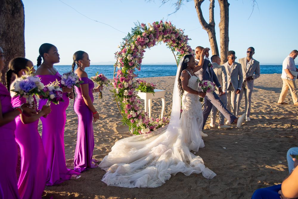 Beach wedding ceremony under a floral arch with bridesmaids in bright purple dresses.