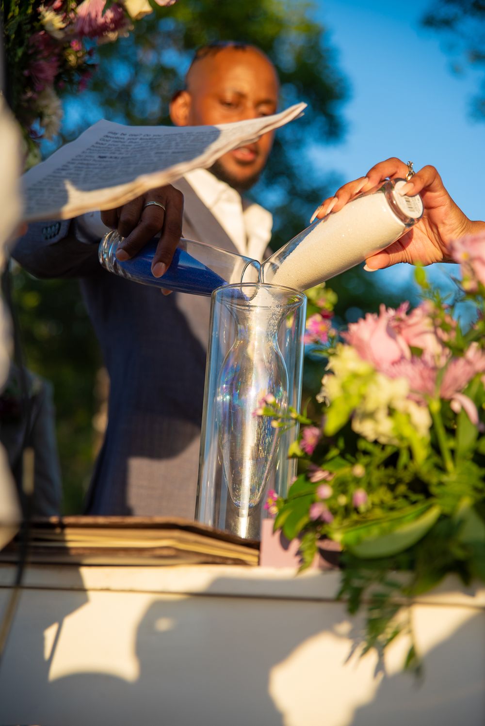Wedding ceremony sand pouring ritual with white and pink floral decorations in outdoor setting.