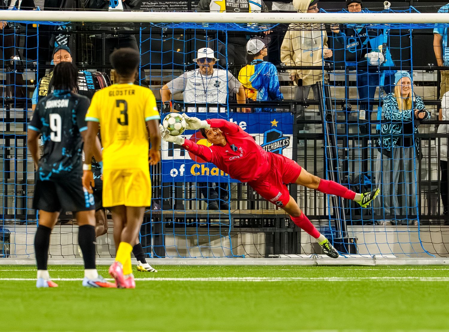 A goalkeeper dives to make a save during a soccer match as spectators watch from the stadium stands.