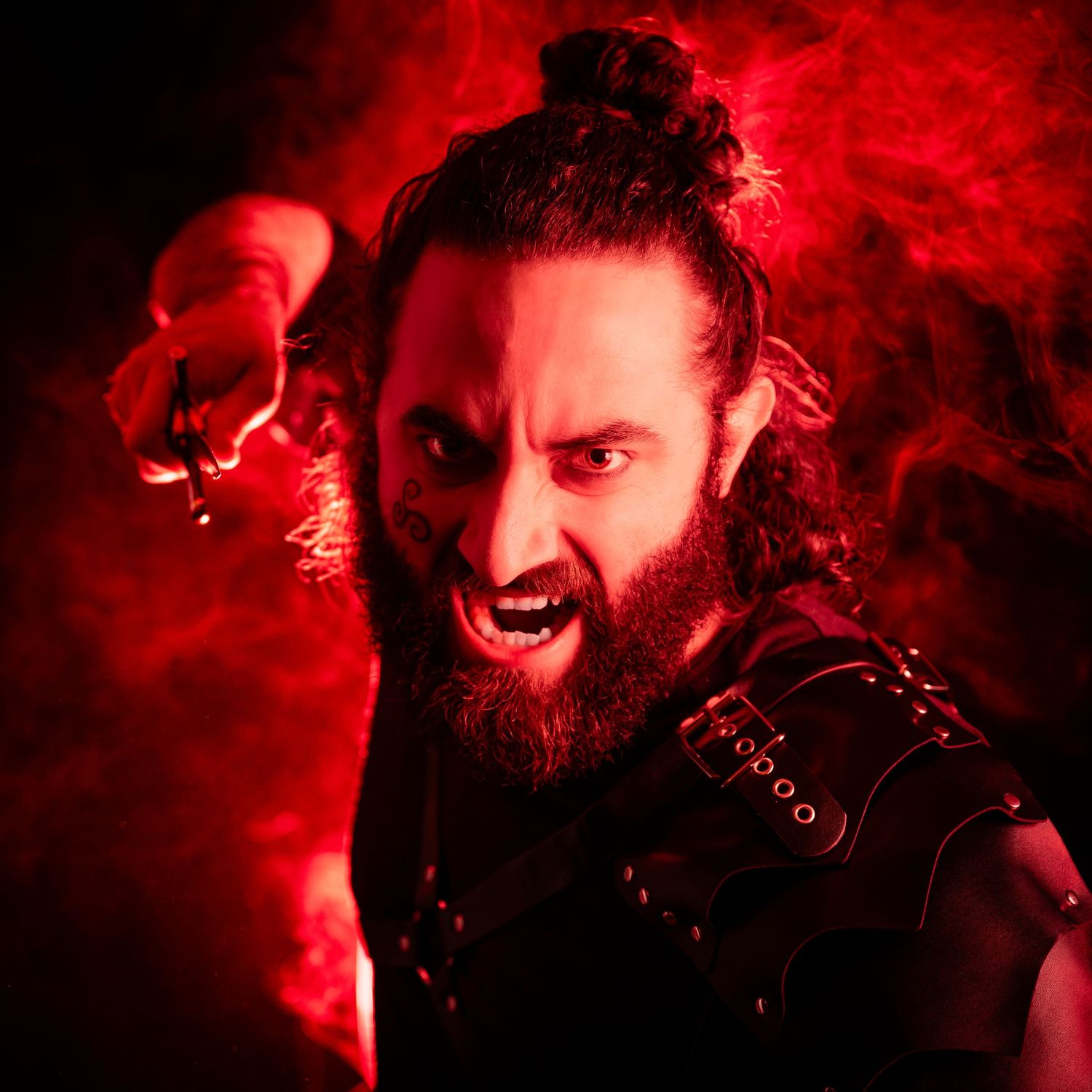 Fierce bearded man in dark armor poses dramatically against red smoky background in fantasy warrior costume.
