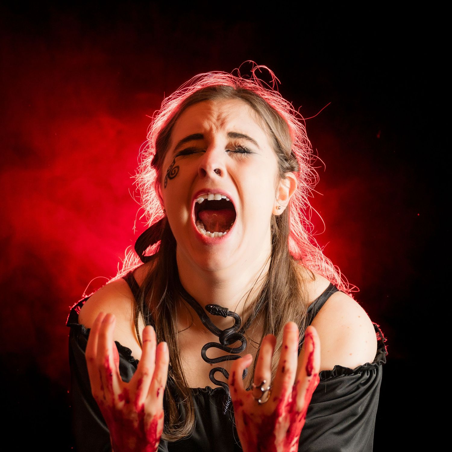 A woman in Halloween vampire costume screams with bloody hands and snake necklace against dramatic red smoky background.
