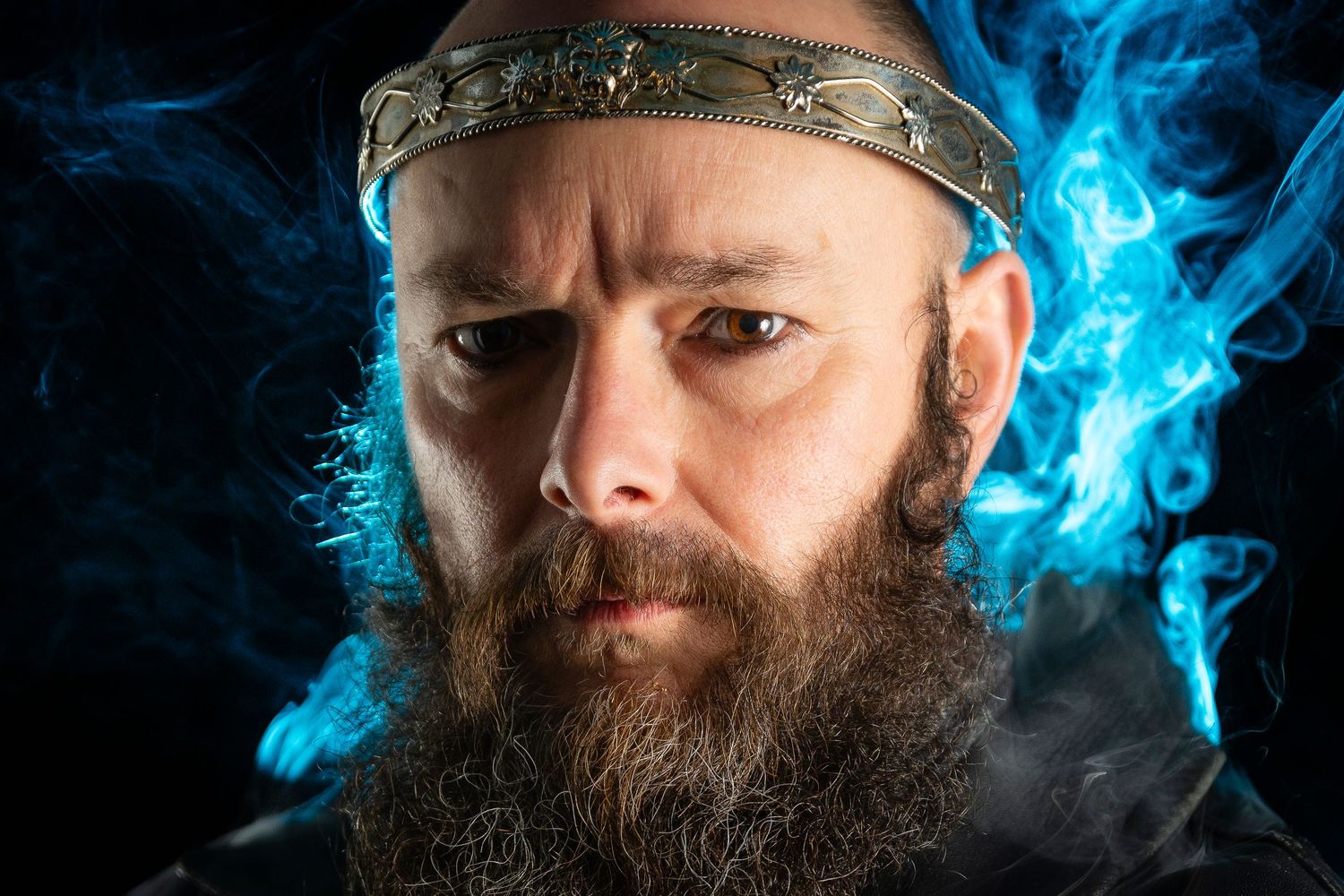 Bearded man wearing a medieval crown with blue smoke swirling around him against a dark background.
