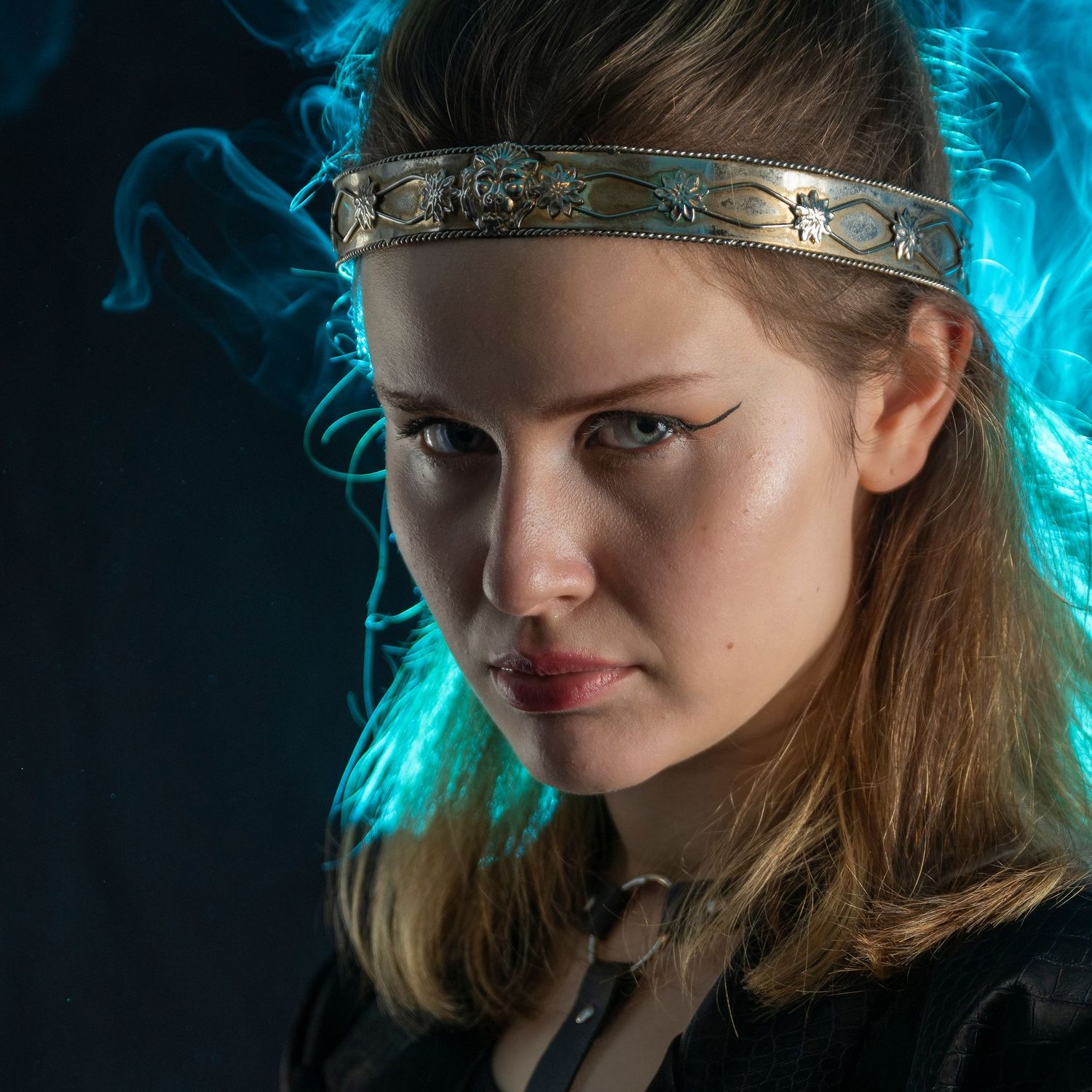 Young woman in medieval costume with ornate headband, dramatic teal smoke background, intense expression.
