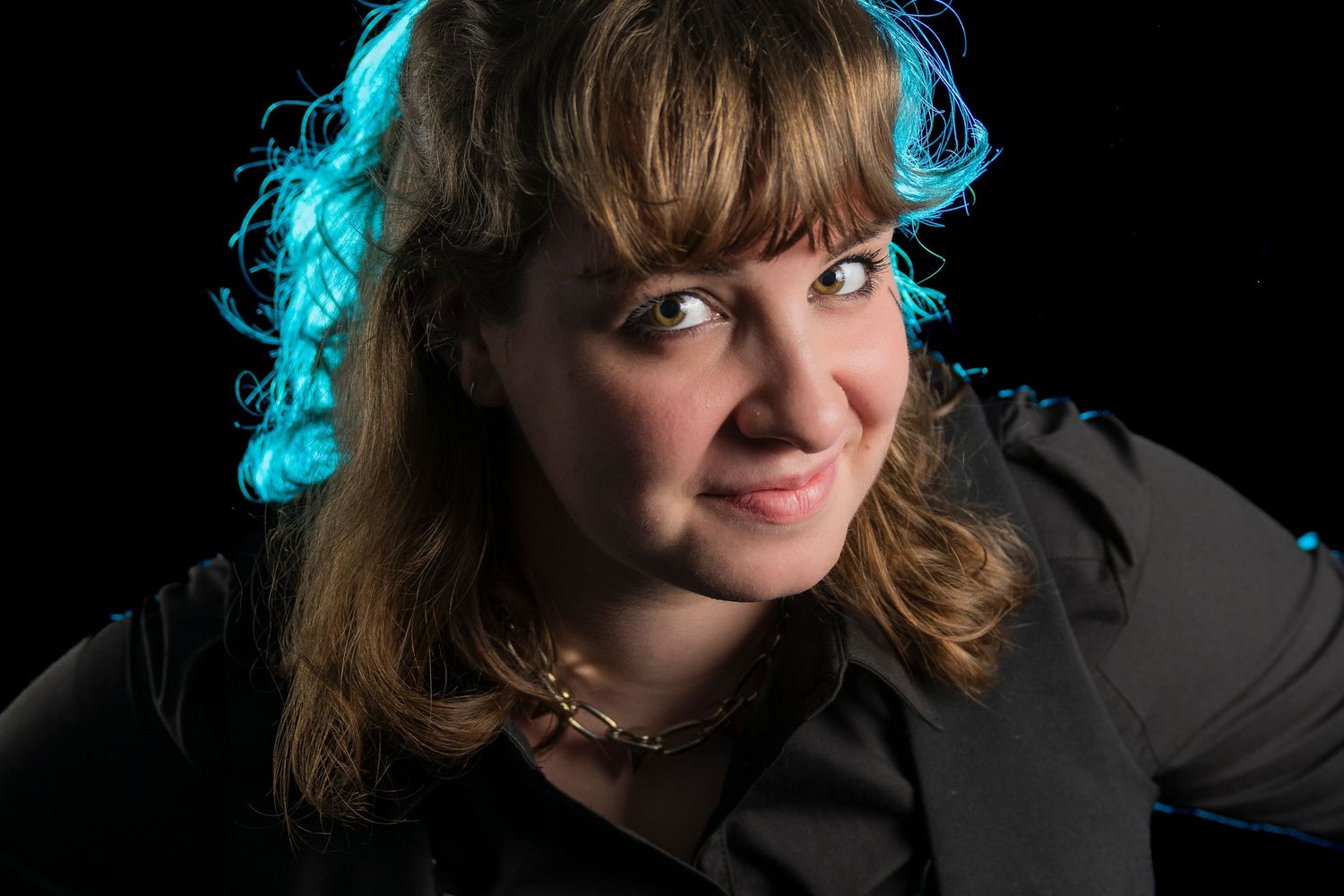 Woman with blonde hair wearing a black shirt and necklace, lit with blue accent lighting against dark background.