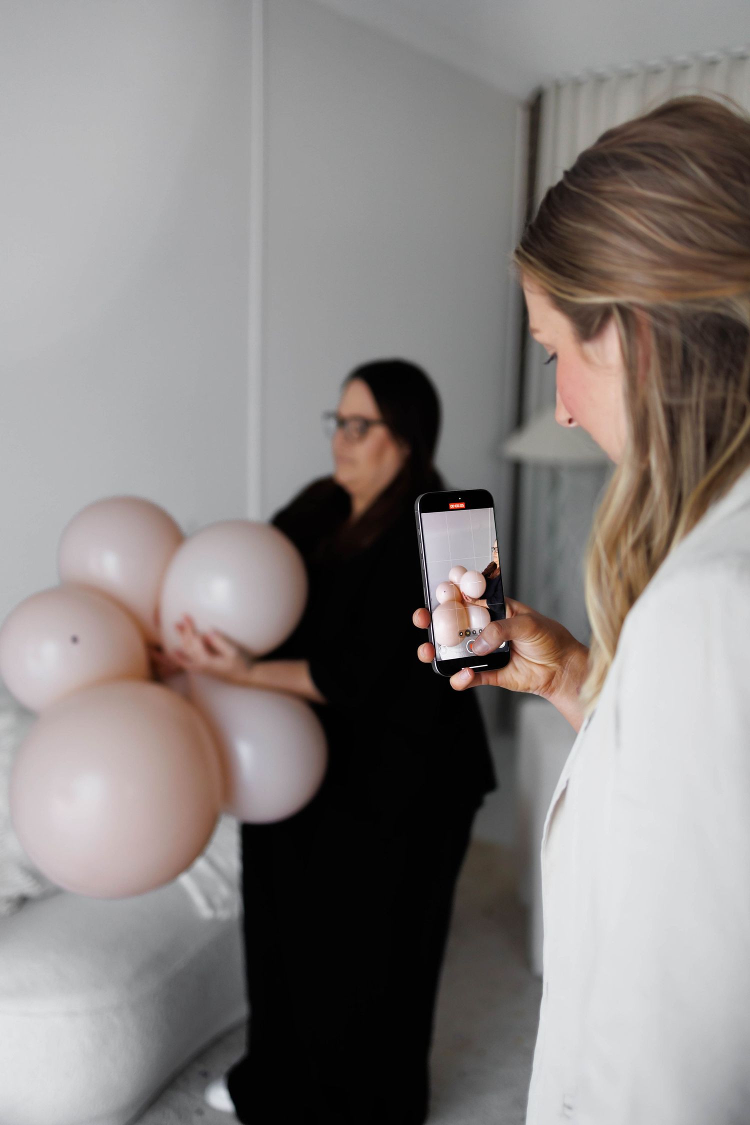 Woman photographs friend holding pink balloons with smartphone in bright modern room.