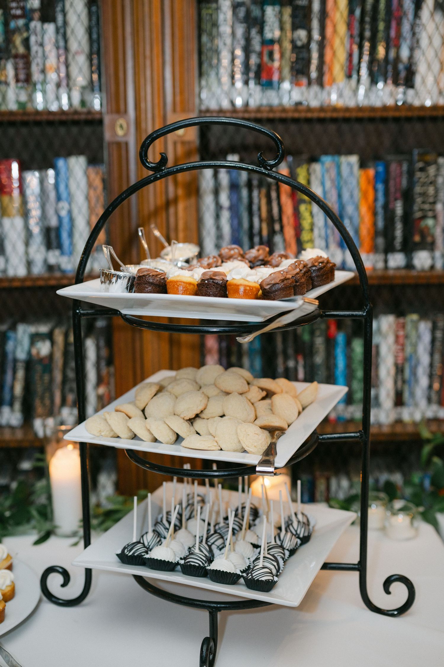 Three-tier black metal dessert stand with cookies, brownies, and cake pops displayed in front of a bookshelf.