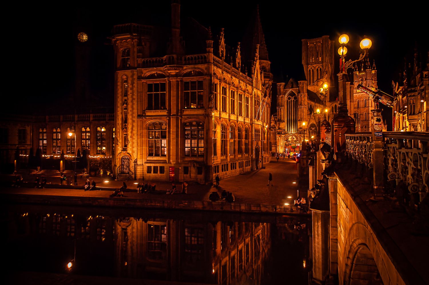 Wide nighttime photo of a brightly lit city square with historic buildings and street lights, reflecting off wet pavement.