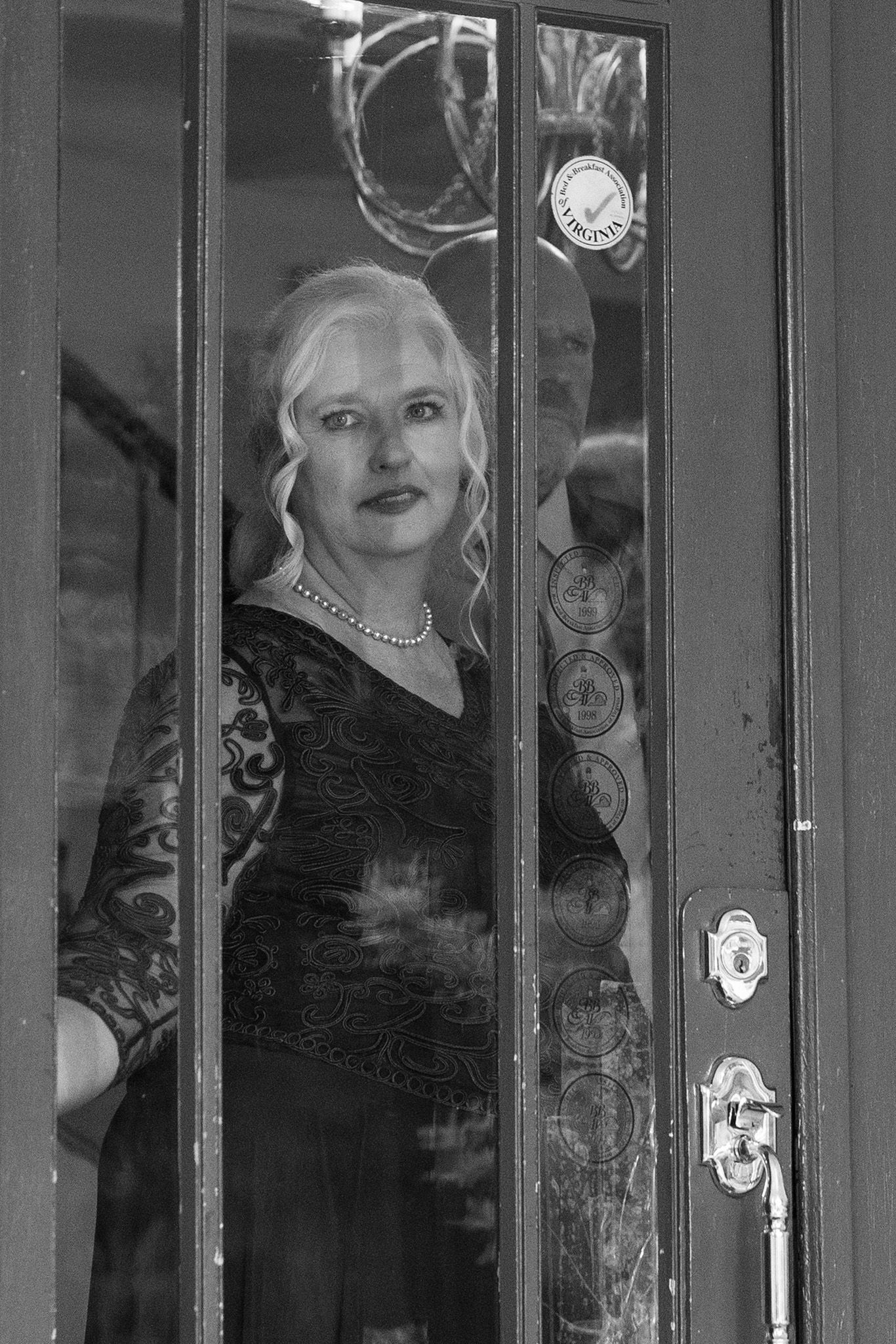 Black and white photo of an elegant blonde woman in lace dress and pearl necklace standing behind a glass door.