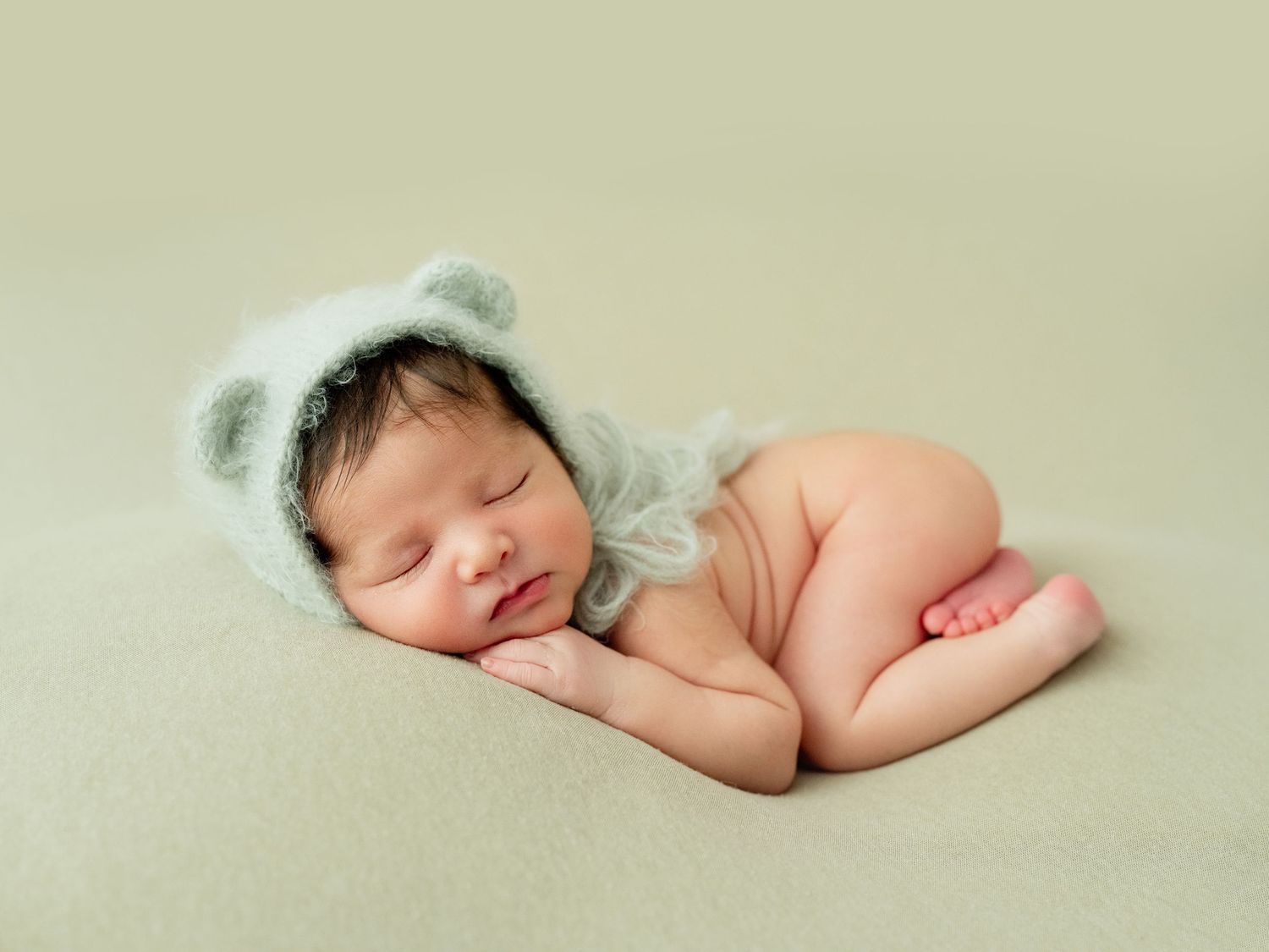Sleeping newborn baby wearing a soft mint green bear bonnet, curled up on a cream blanket in a peaceful pose.