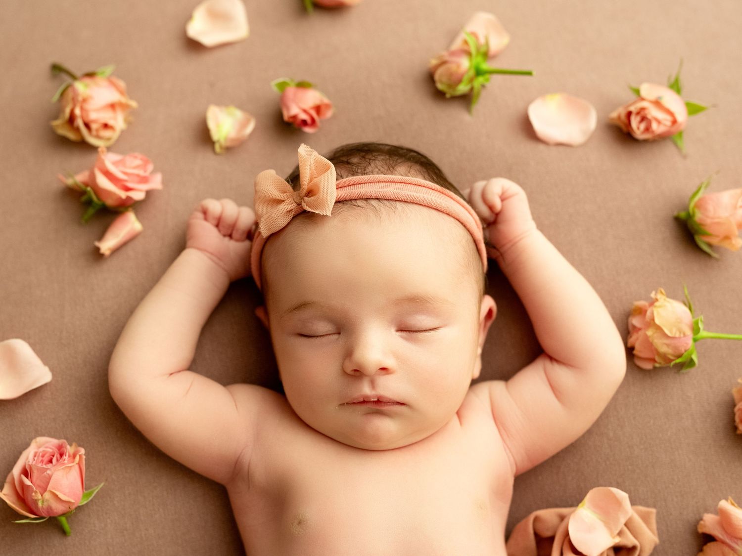 Sleeping newborn baby girl with pink bow headband surrounded by soft peach roses on brown background.