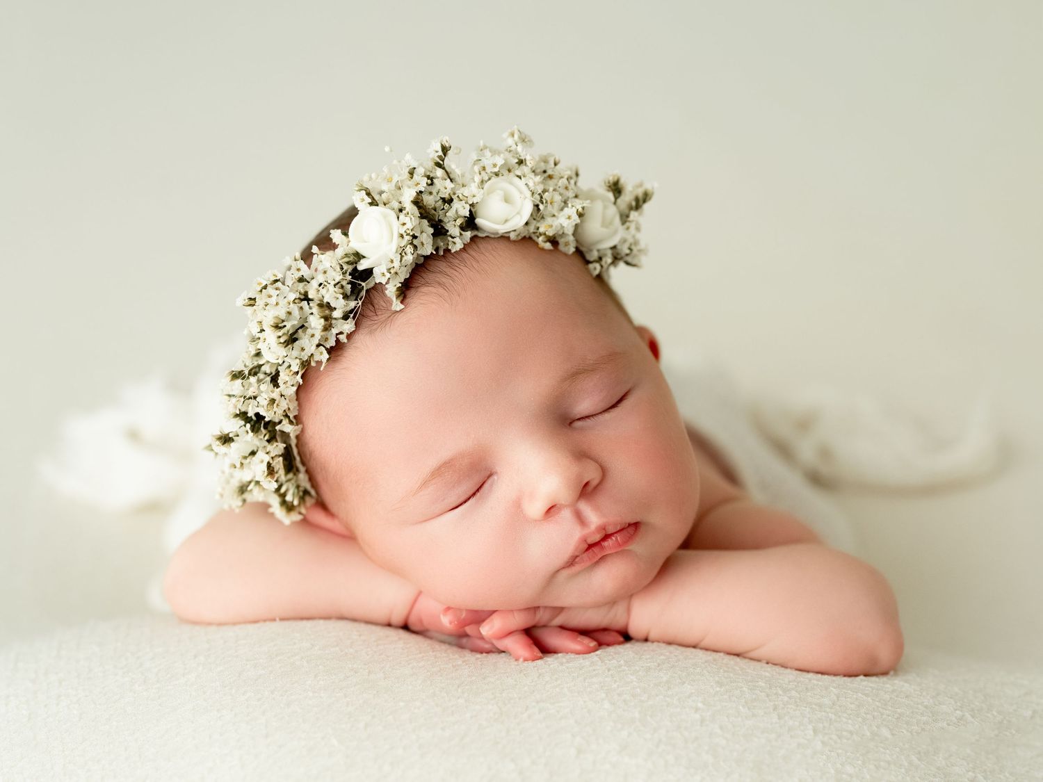 Sleeping newborn baby wearing a delicate floral crown, resting chin on hands on a soft white blanket.