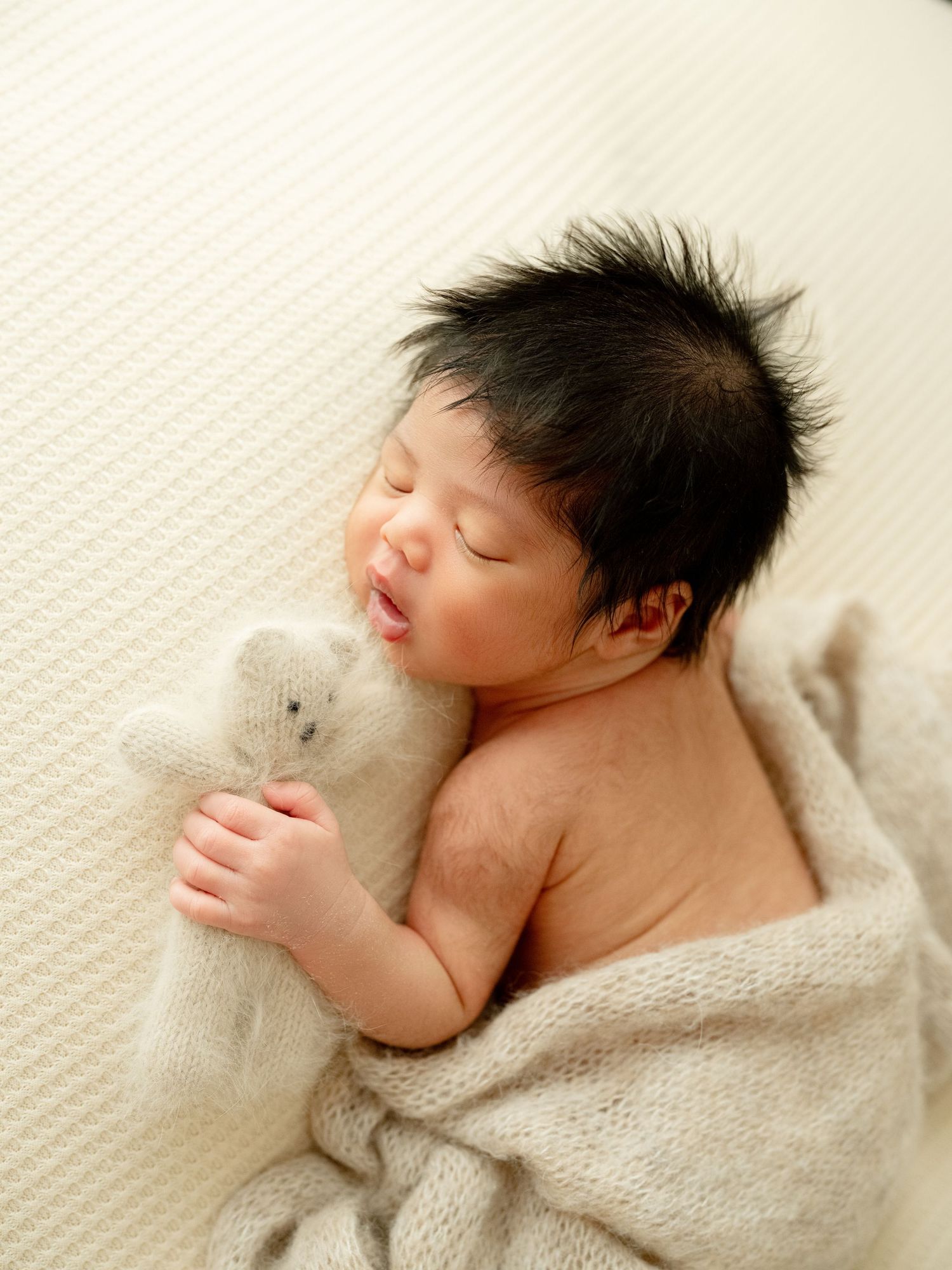 Sleeping newborn baby with dark hair cuddling a small teddy bear, wrapped in a soft knit blanket.