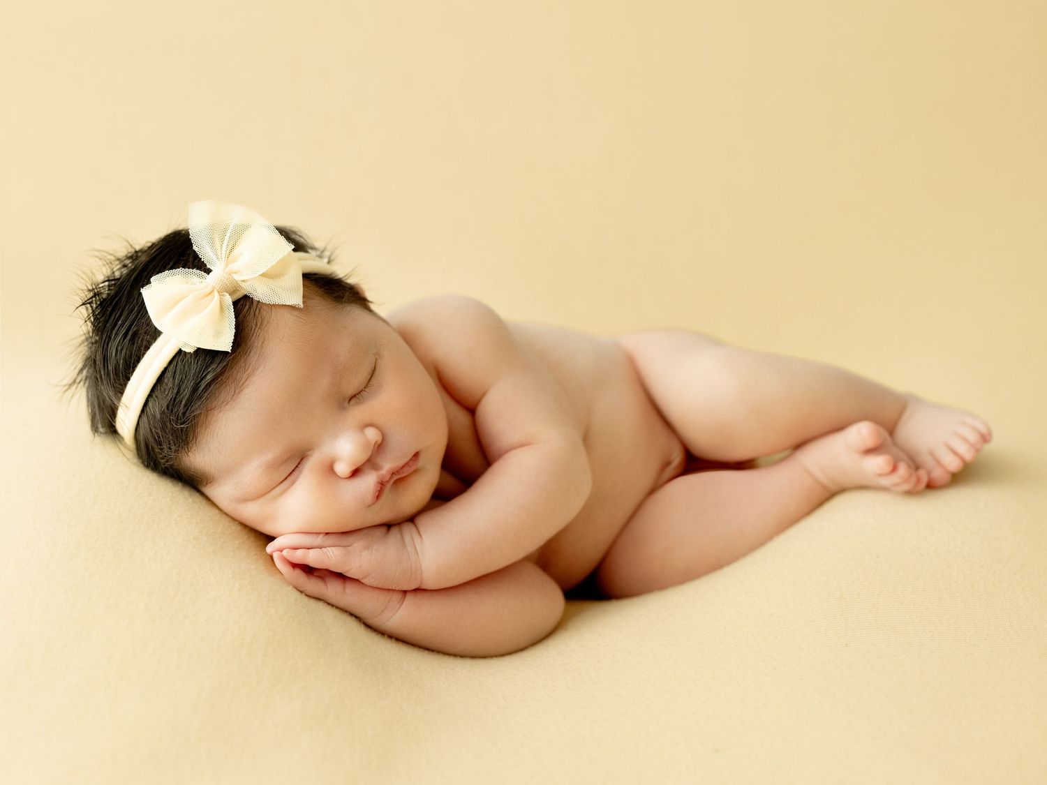 Newborn baby sleeping peacefully on yellow backdrop, wearing cream bow headband, posed in froggy position.
