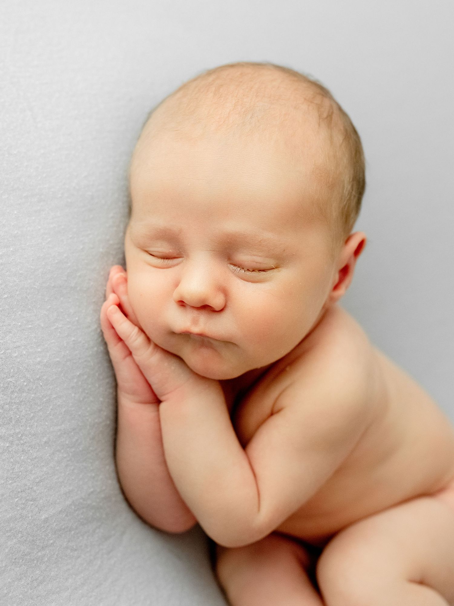 Peaceful newborn baby sleeping curled up with hands tucked under cheek on soft gray background.