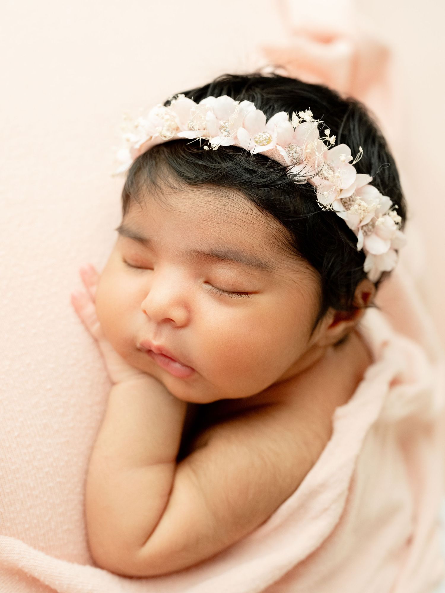 Sleeping newborn baby girl wearing a delicate pink floral headband, wrapped in a soft pink blanket.