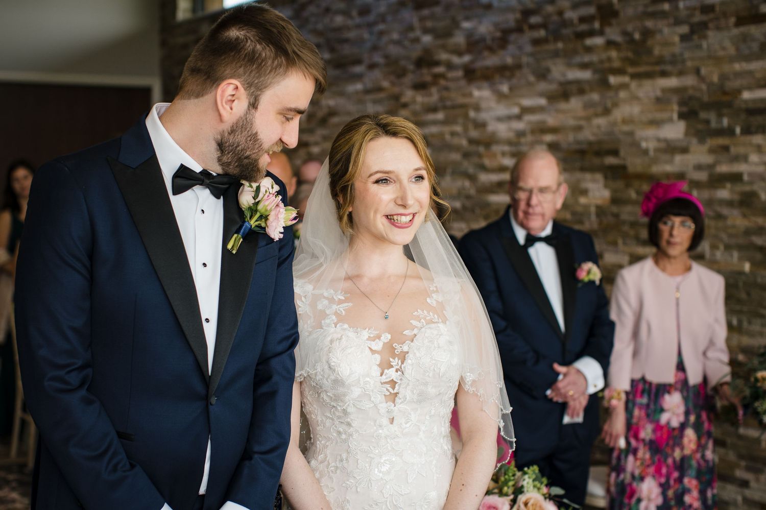 Bride and groom smiling at wedding ceremony, bride in lace gown, groom in navy tuxedo, guests in background.