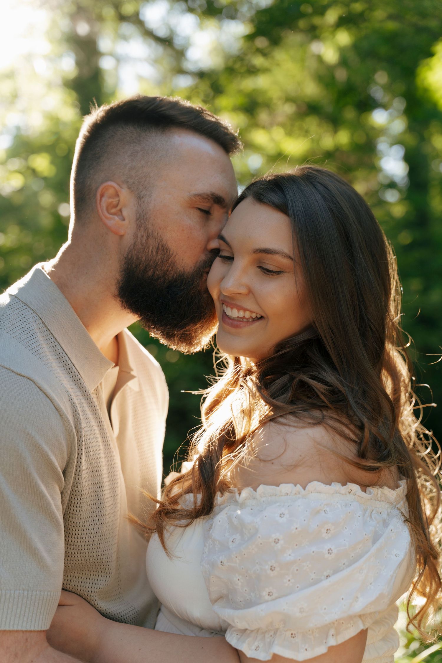 A bearded man in a tan shirt embraces a woman in a white off-shoulder dress, standing in a sunlit garden.