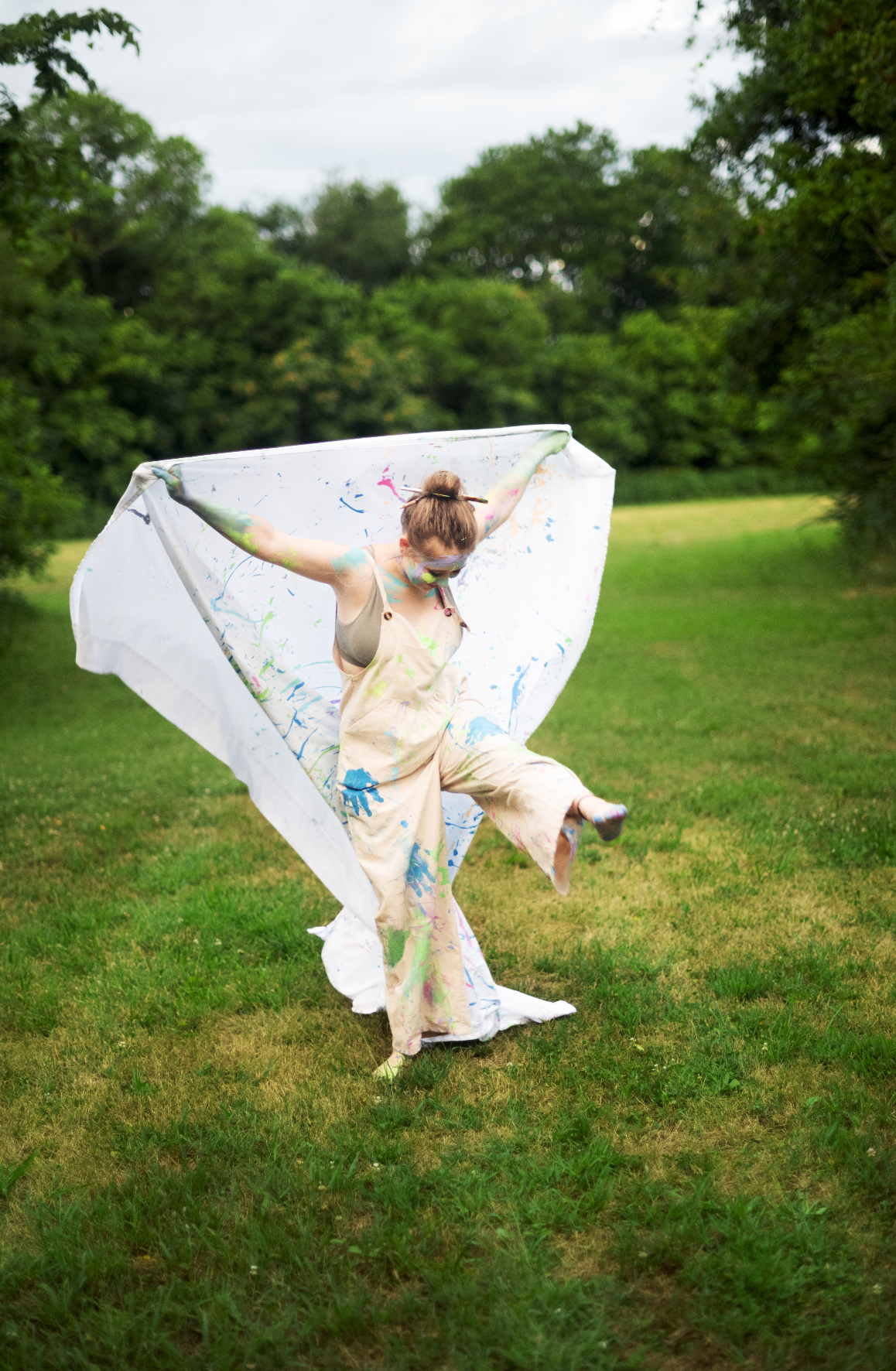 Artist in paint-splattered overalls twirls a white canvas sheet outdoors in a lush green park.