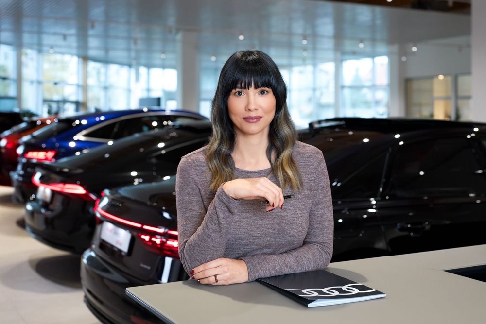 Woman leaning on a desk in a modern car dealership showroom with luxury black vehicles displayed behind her.