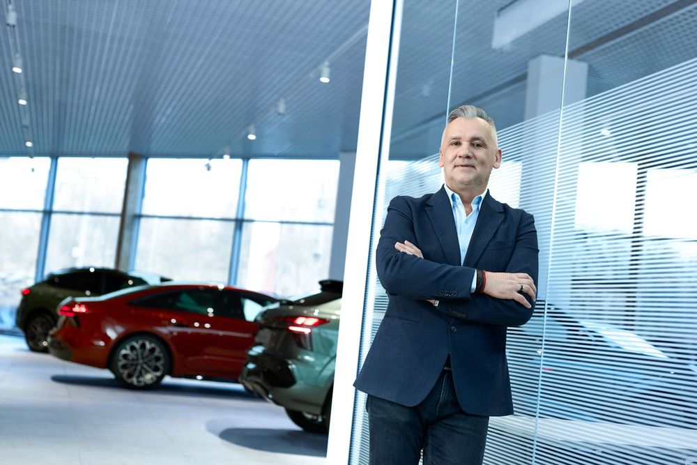 Confident businessman in a suit stands arms crossed in a modern car showroom with luxury vehicles in the background.