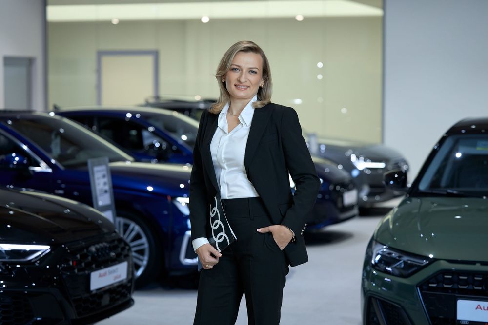 Professional woman in black suit poses confidently in modern Audi car dealership showroom surrounded by luxury vehicles.