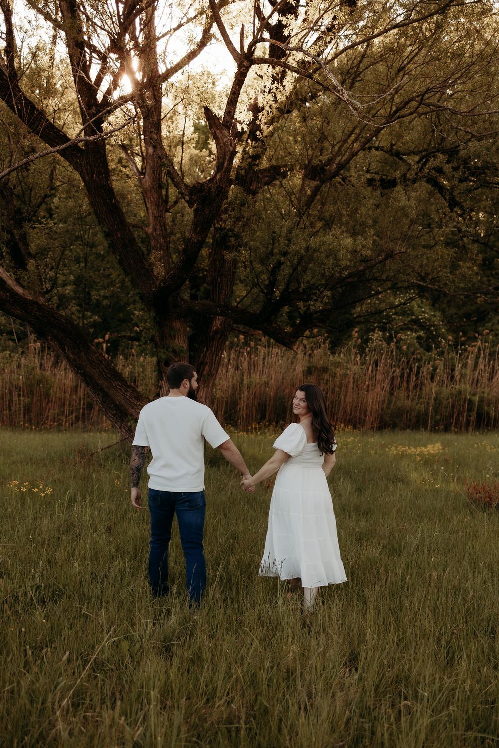 Couple holding hands in a sunlit meadow, woman in white dress and man in white shirt and jeans, surrounded by tall grass and trees.