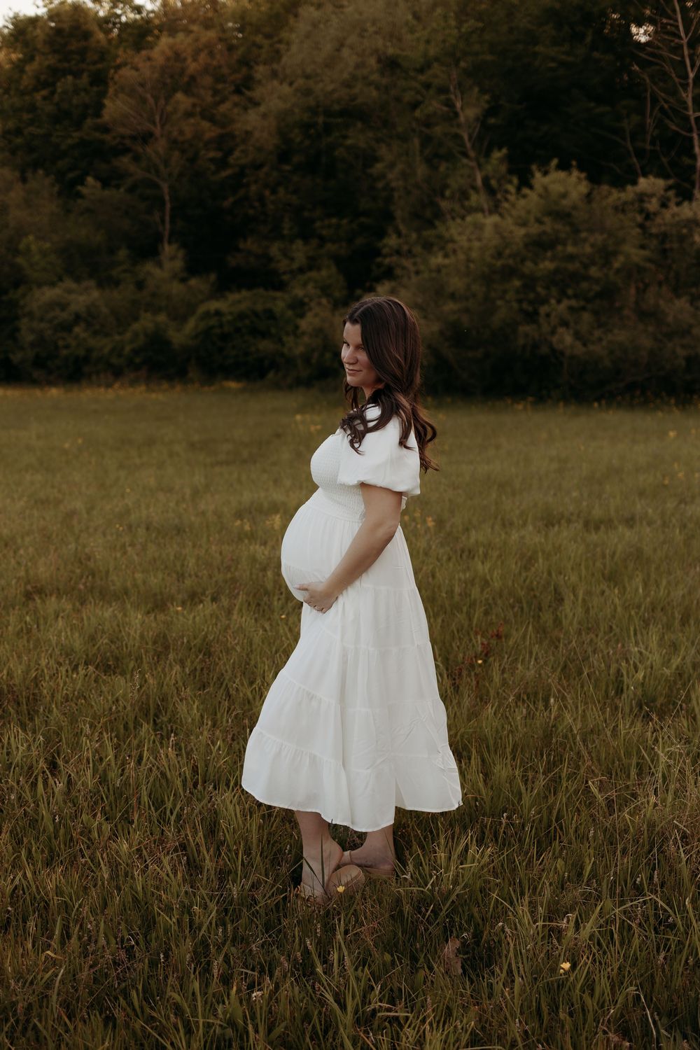Pregnant woman in white off-shoulder dress standing in green field at golden hour, cradling baby bump.