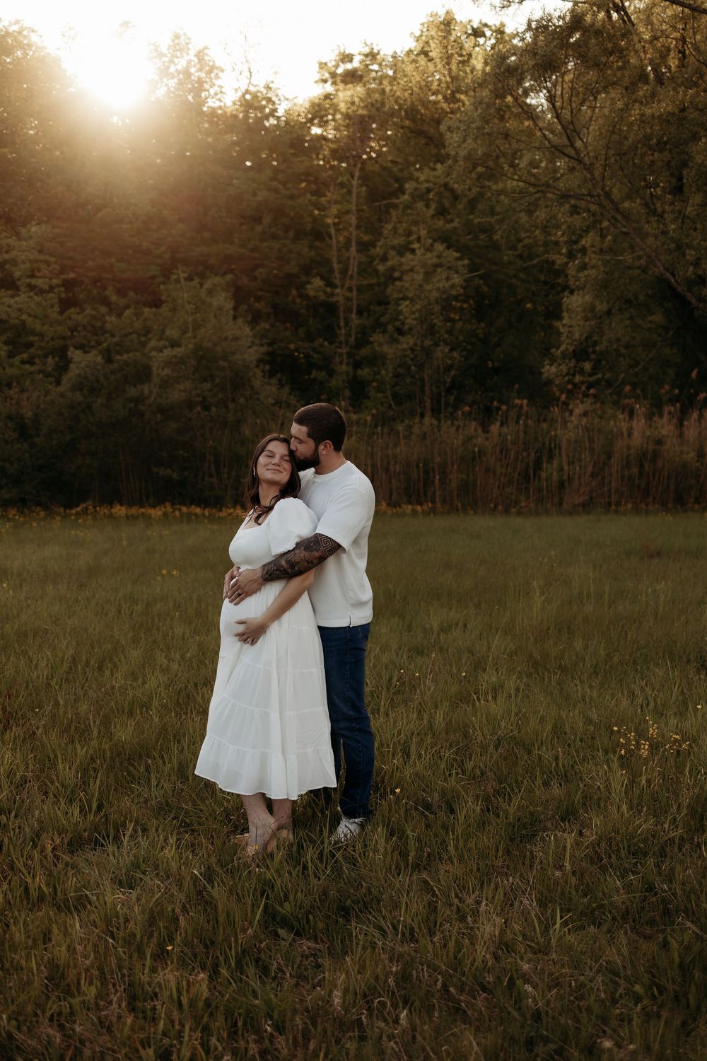 A couple embraces in a sunlit meadow at golden hour, the woman in a white dress cradling her baby bump.
