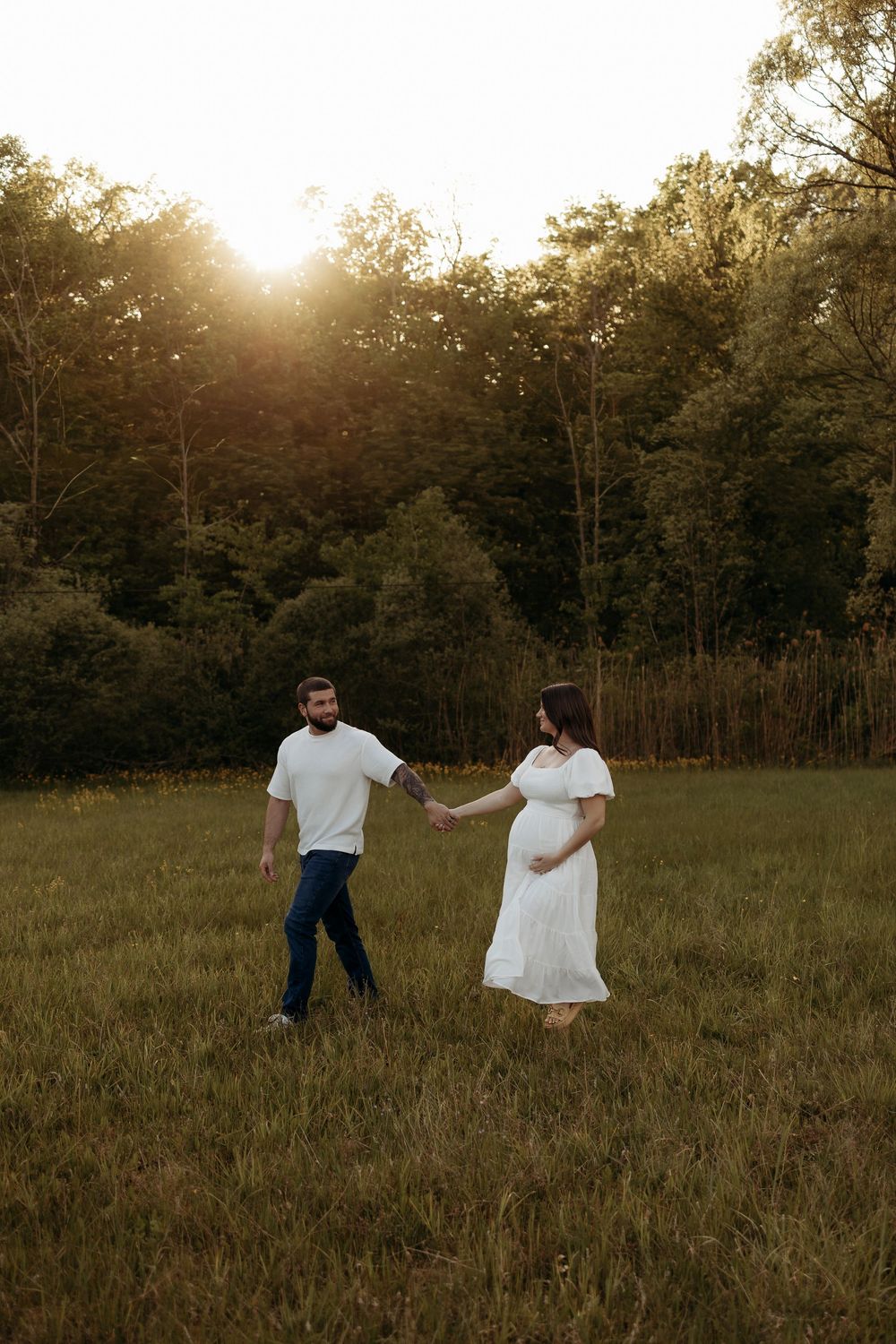Pregnant couple holding hands walking in a sunlit meadow at golden hour, woman in white dress, man in white shirt and jeans.