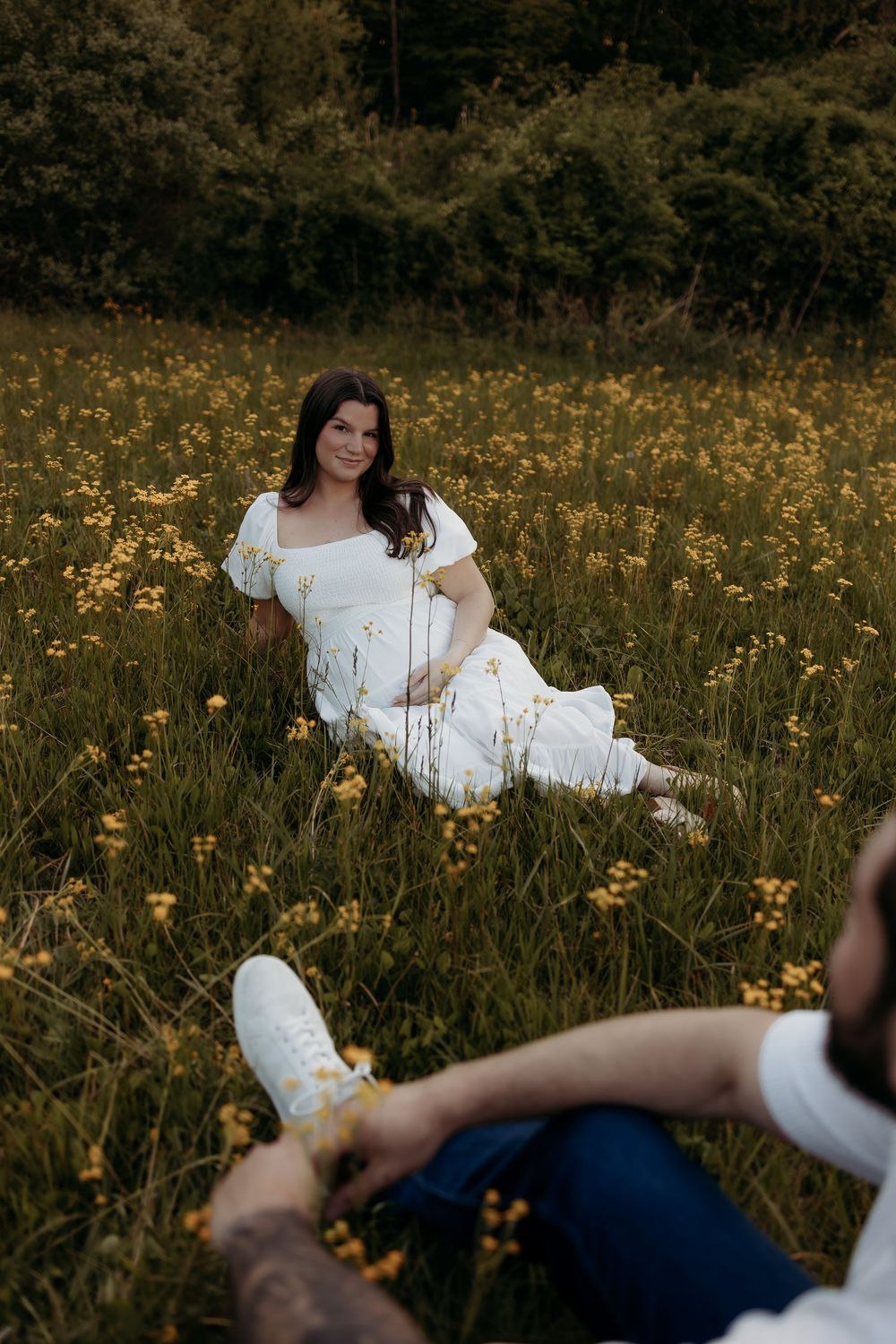 Pregnant woman in white dress sitting in yellow wildflower field, smiling at partner during outdoor maternity photoshoot.