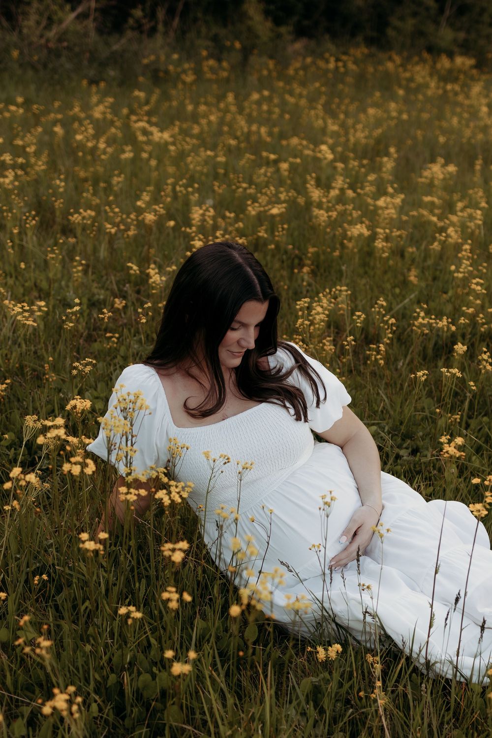Pregnant woman in white dress sitting in a field of yellow wildflowers, gently cradling her baby bump.