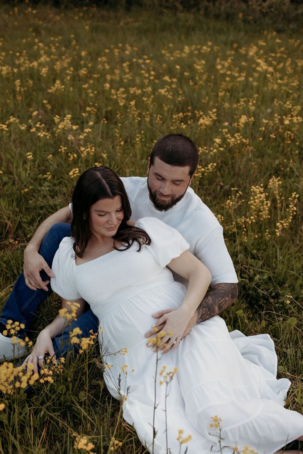 Pregnant couple sitting in a yellow wildflower field, man embracing woman in white maternity dress.