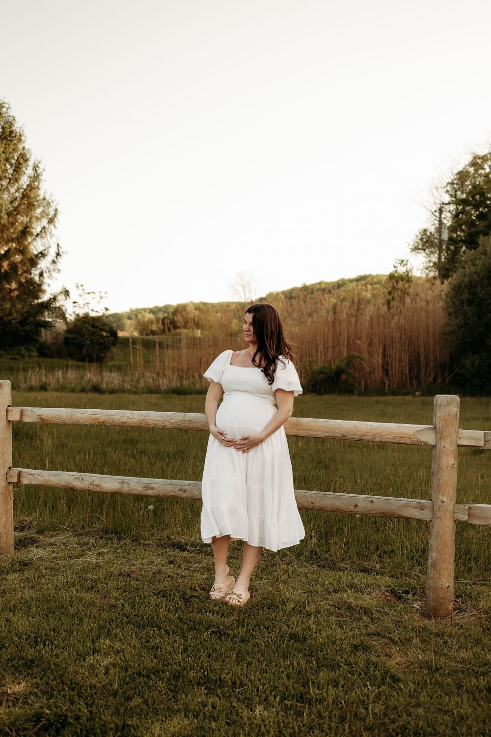 Pregnant woman in white dress holding baby bump by rustic fence in golden hour field.