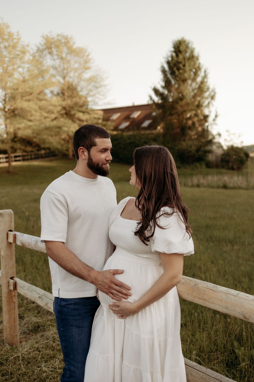 A couple in white outfits cradles a pregnant belly near a wooden fence in a golden hour outdoor maternity photo.