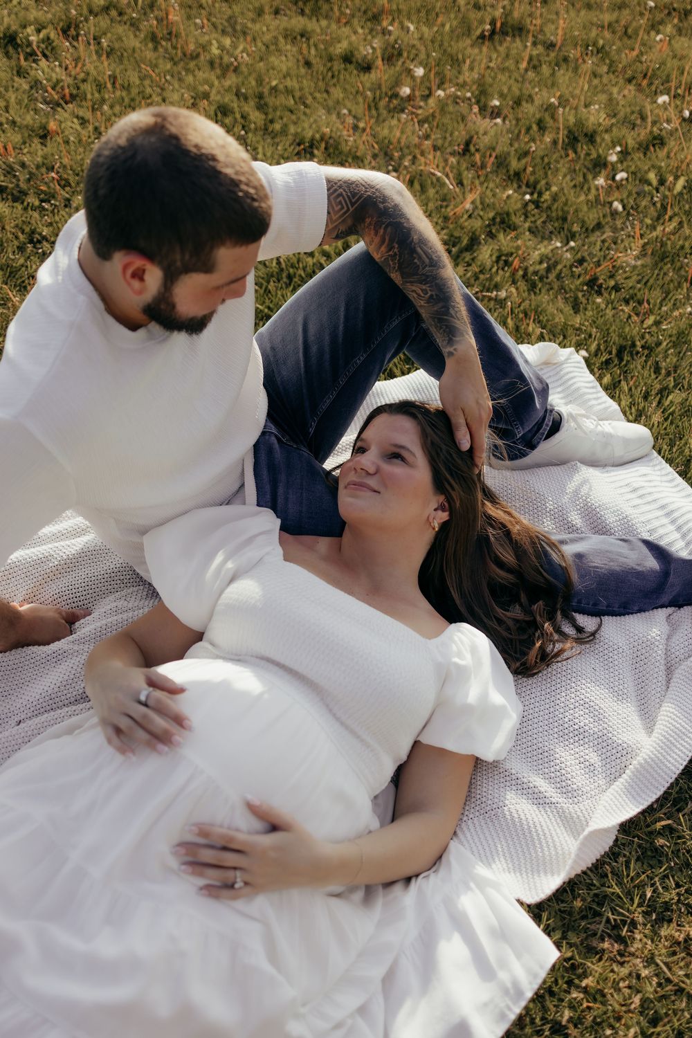 Pregnant woman in white dress lying on blanket smiling up at bearded tattooed partner outdoors.