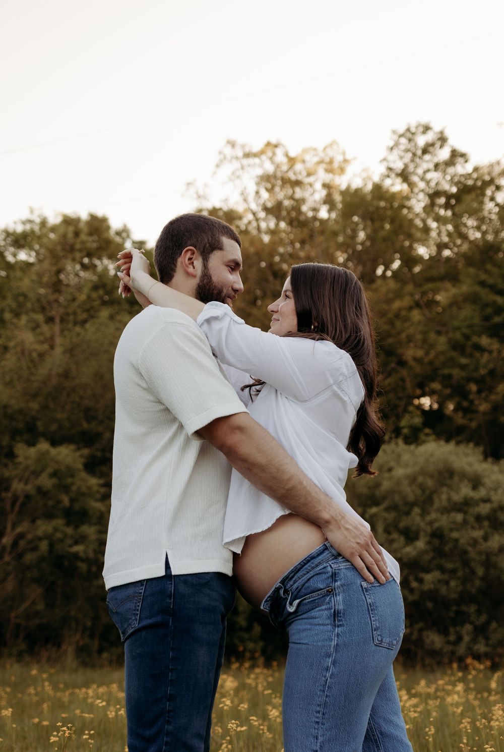 Couple embracing in a golden field, woman showing baby bump, surrounded by lush green trees at sunset.