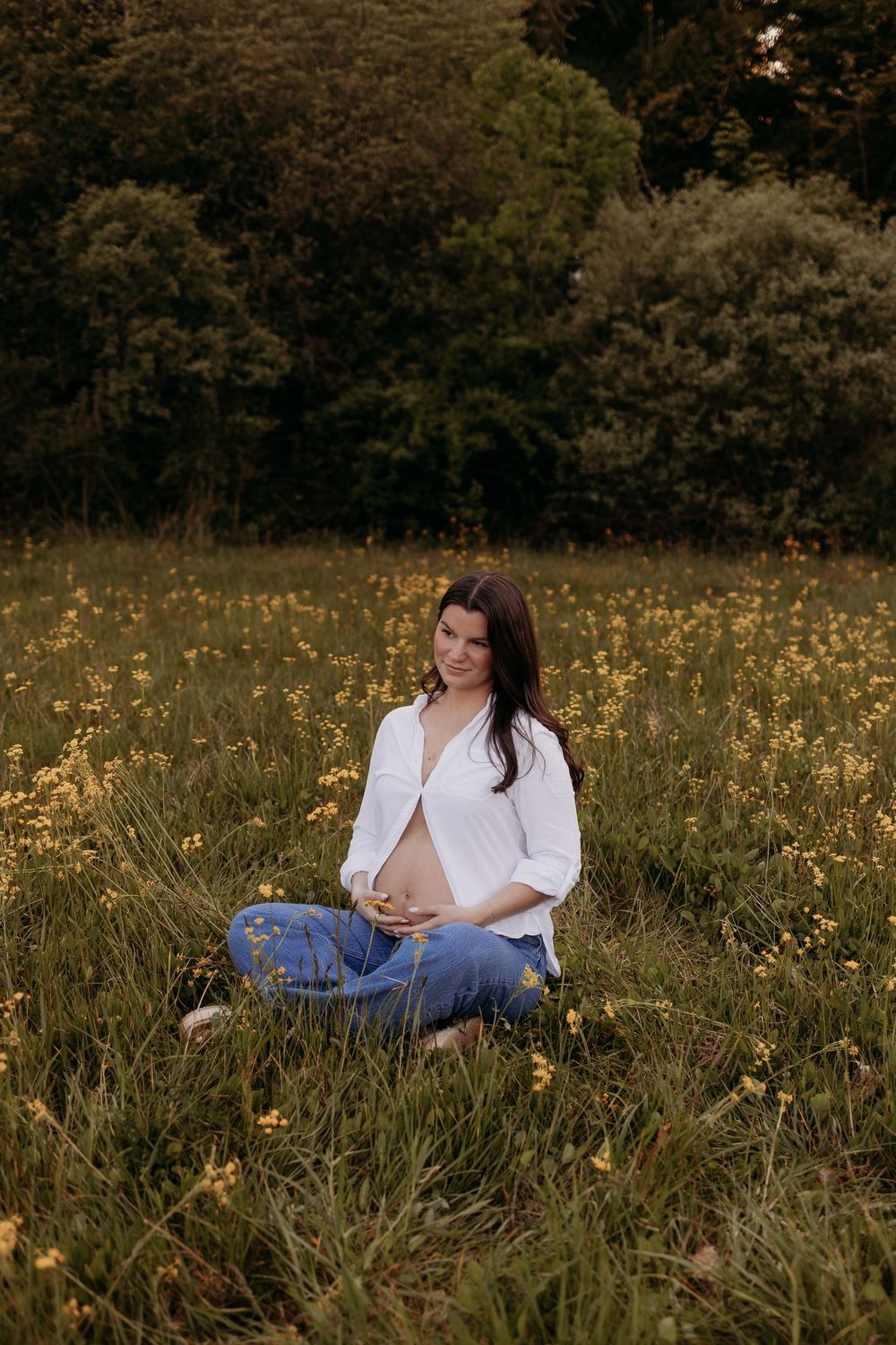 Pregnant woman sitting cross-legged in a wildflower meadow, wearing white shirt and jeans, cradling her baby bump.