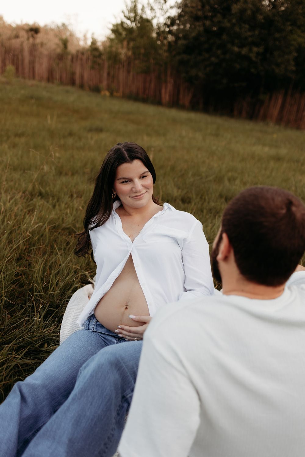 Couple sitting in grass field at sunset, pregnant woman in white shirt cradling baby bump.