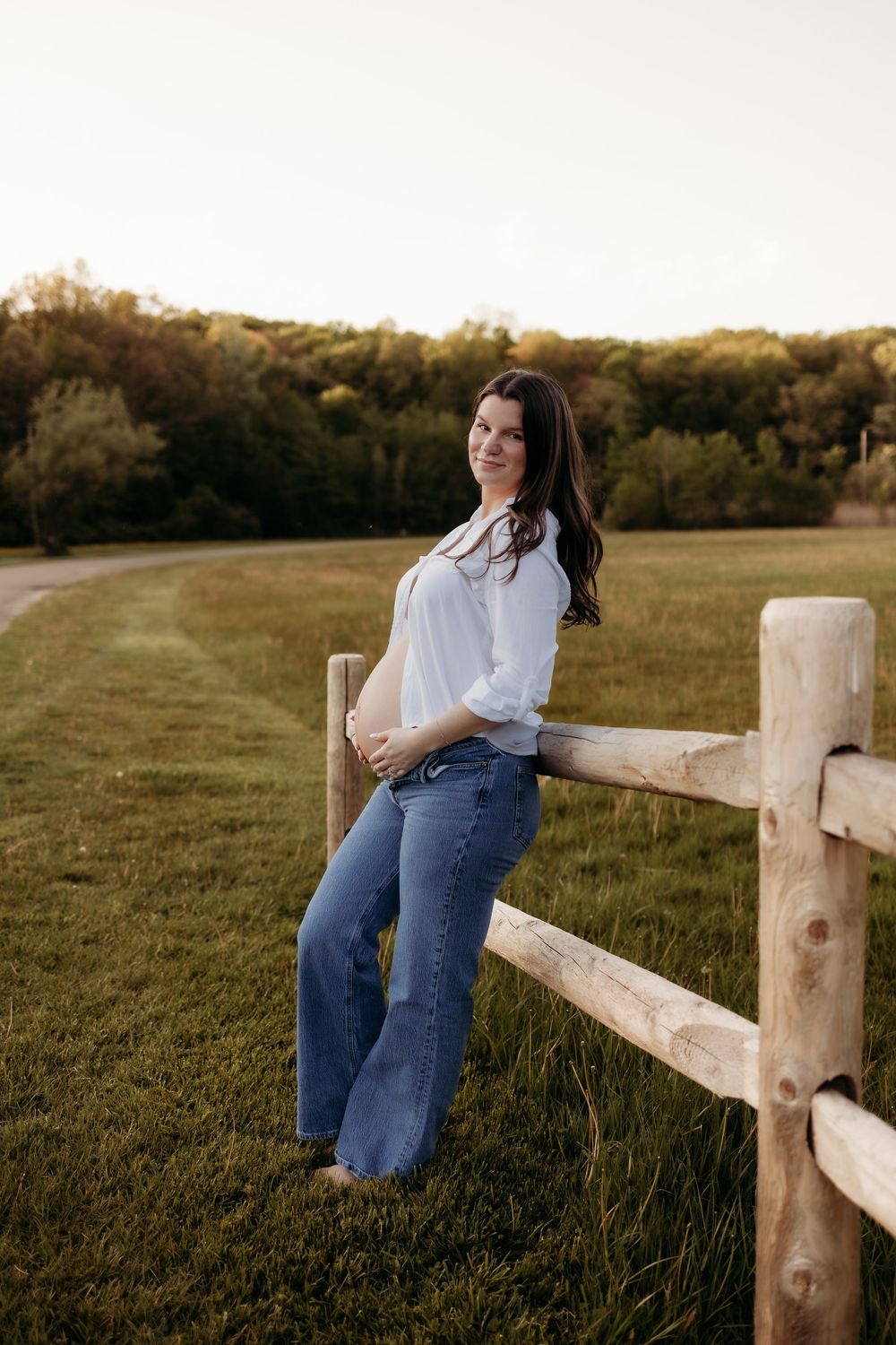 A smiling brunette woman in a white shirt and jeans leans against a wooden fence in a green countryside field.