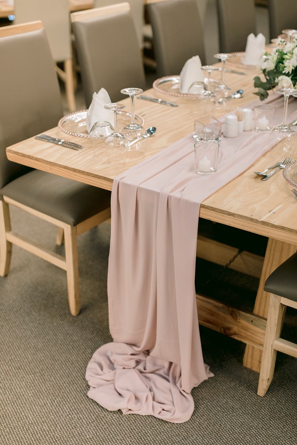 Elegant wedding table setting with blush pink chiffon runner draped over rustic wood table with glassware and white napkins.