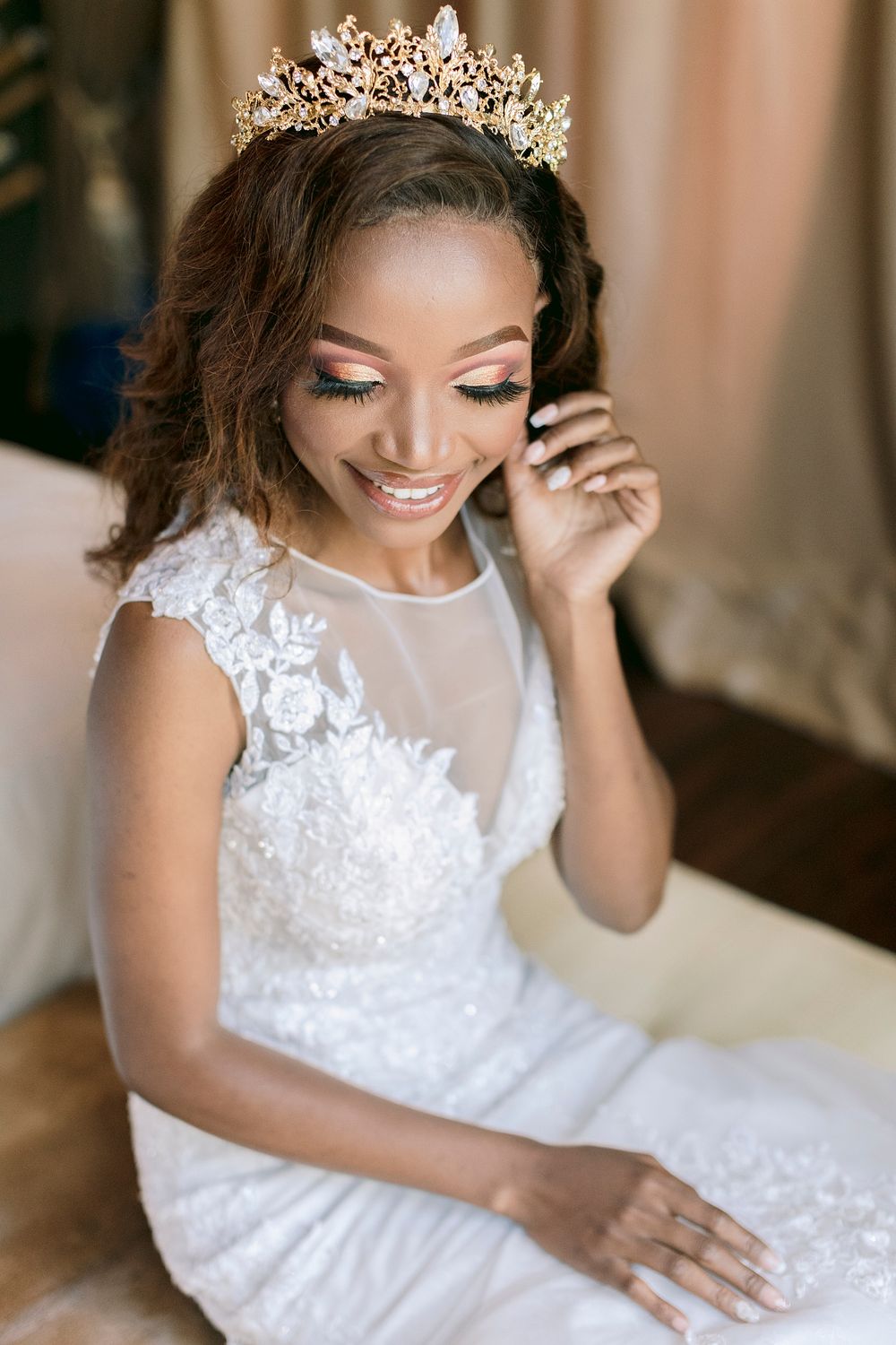 Radiant Black bride wearing white lace wedding gown and ornate gold crown tiara smiling beautifully while seated.