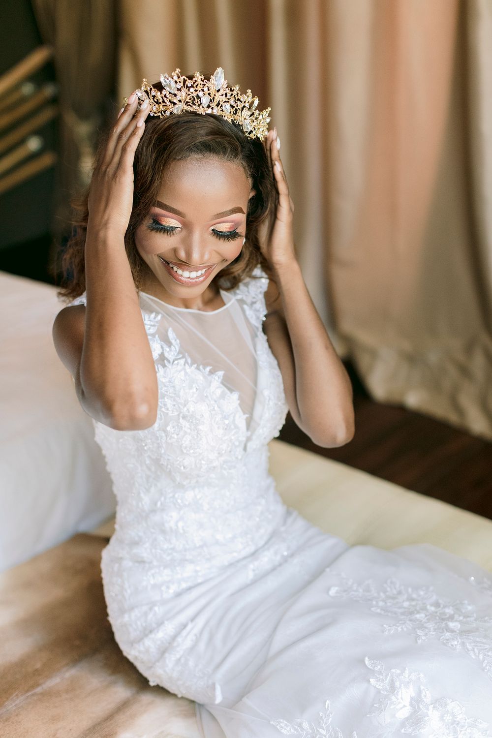 Smiling Black bride in white lace gown adjusting gold crown tiara while seated, wearing elegant bridal makeup.