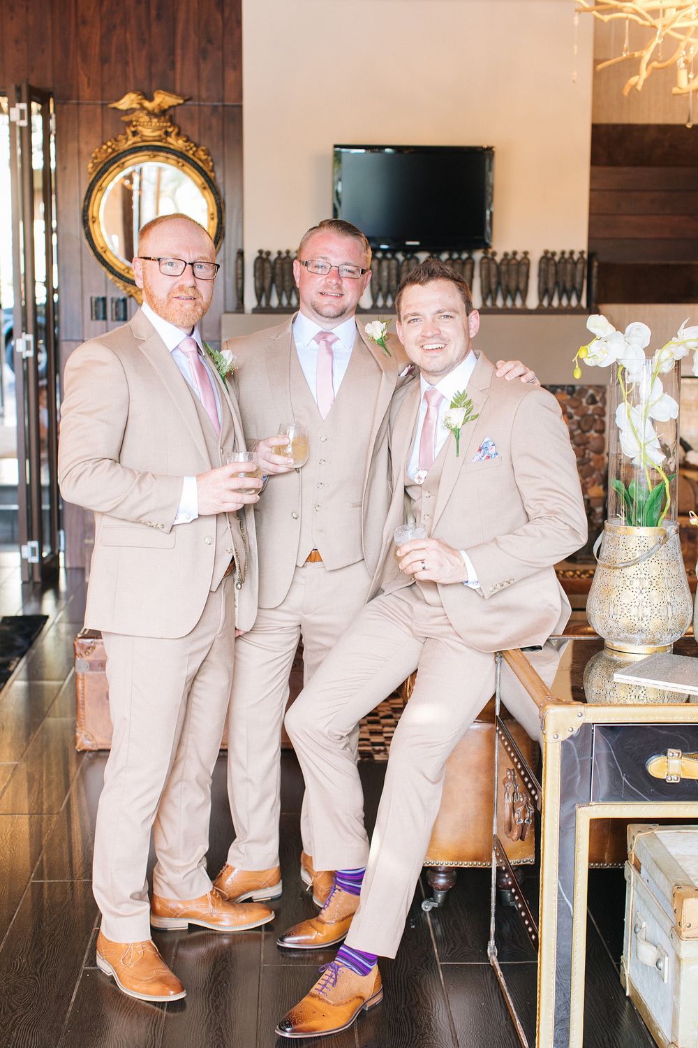 Three groomsmen in tan suits and brown leather shoes smile and hold drinks at a wedding venue.