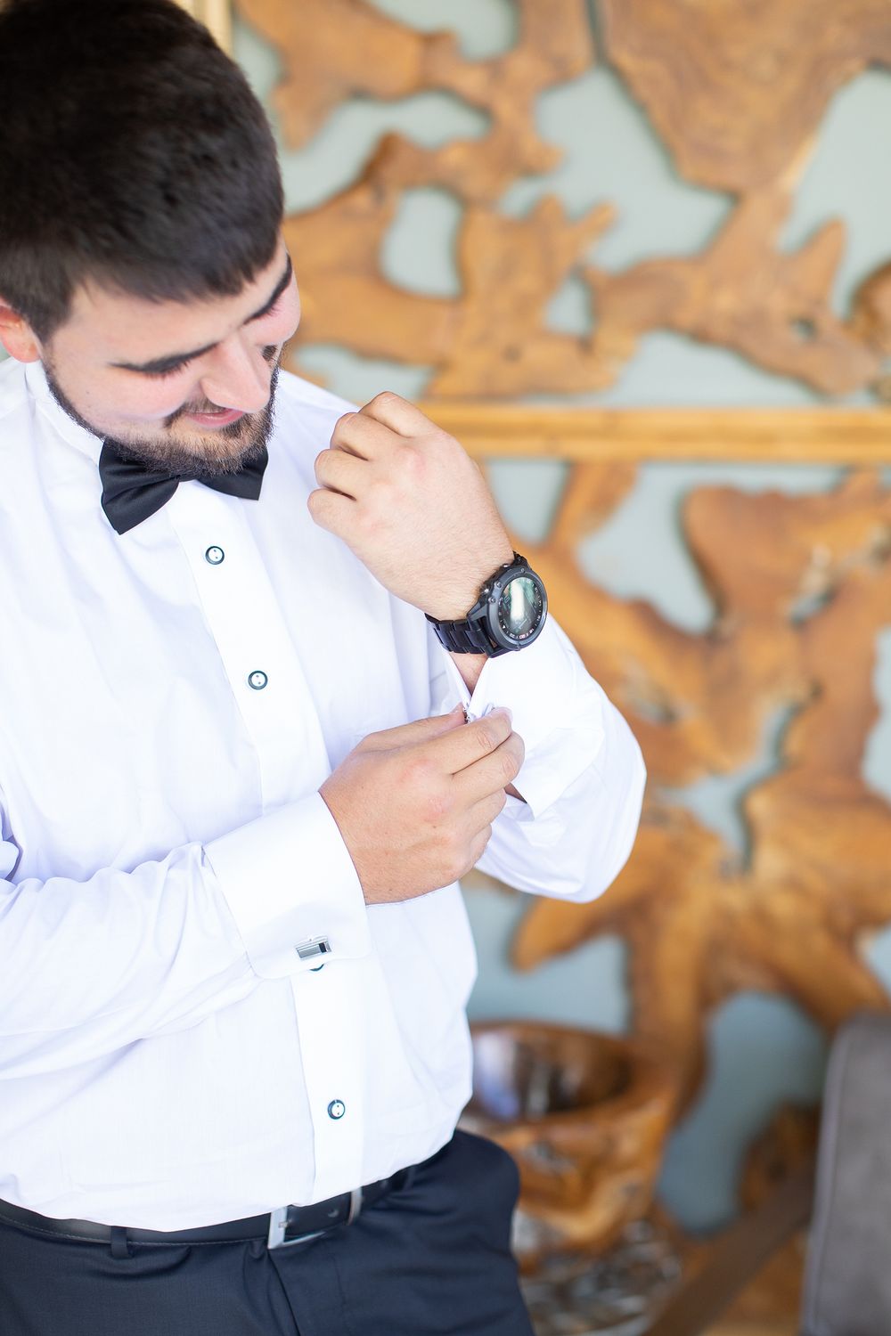 Groom in white shirt and bow tie buttoning his cuff while wearing a black watch before wedding ceremony.