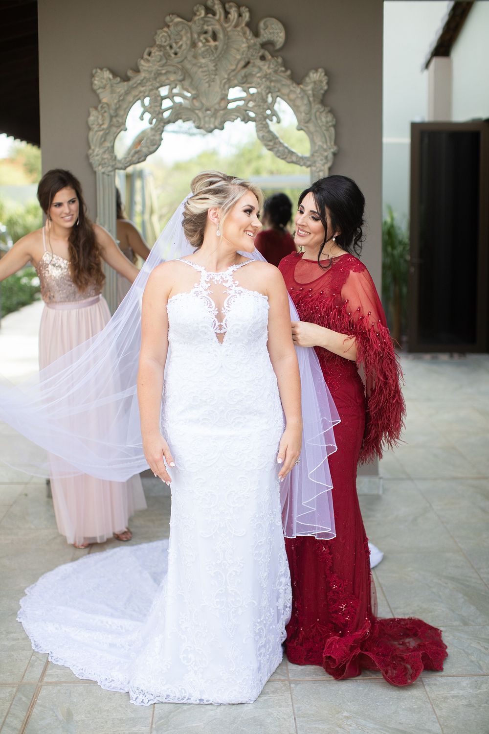 Bride in white lace gown with veil, flanked by bridesmaids in pink and red dresses, posing before ornate mirror.