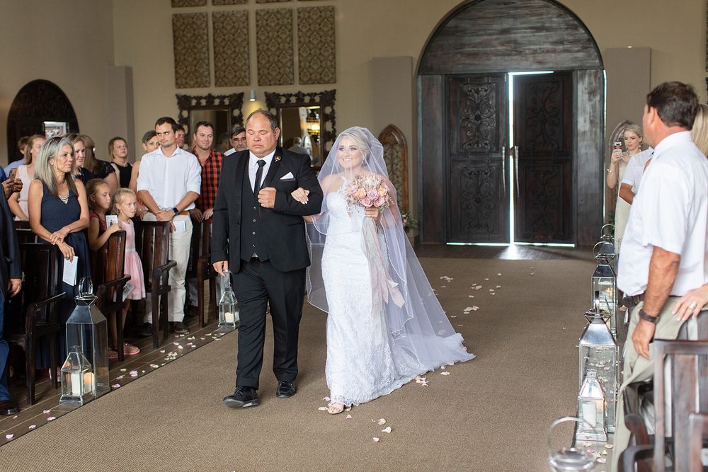 Bride in white gown and veil walks down the aisle with escort at an elegant indoor wedding ceremony.