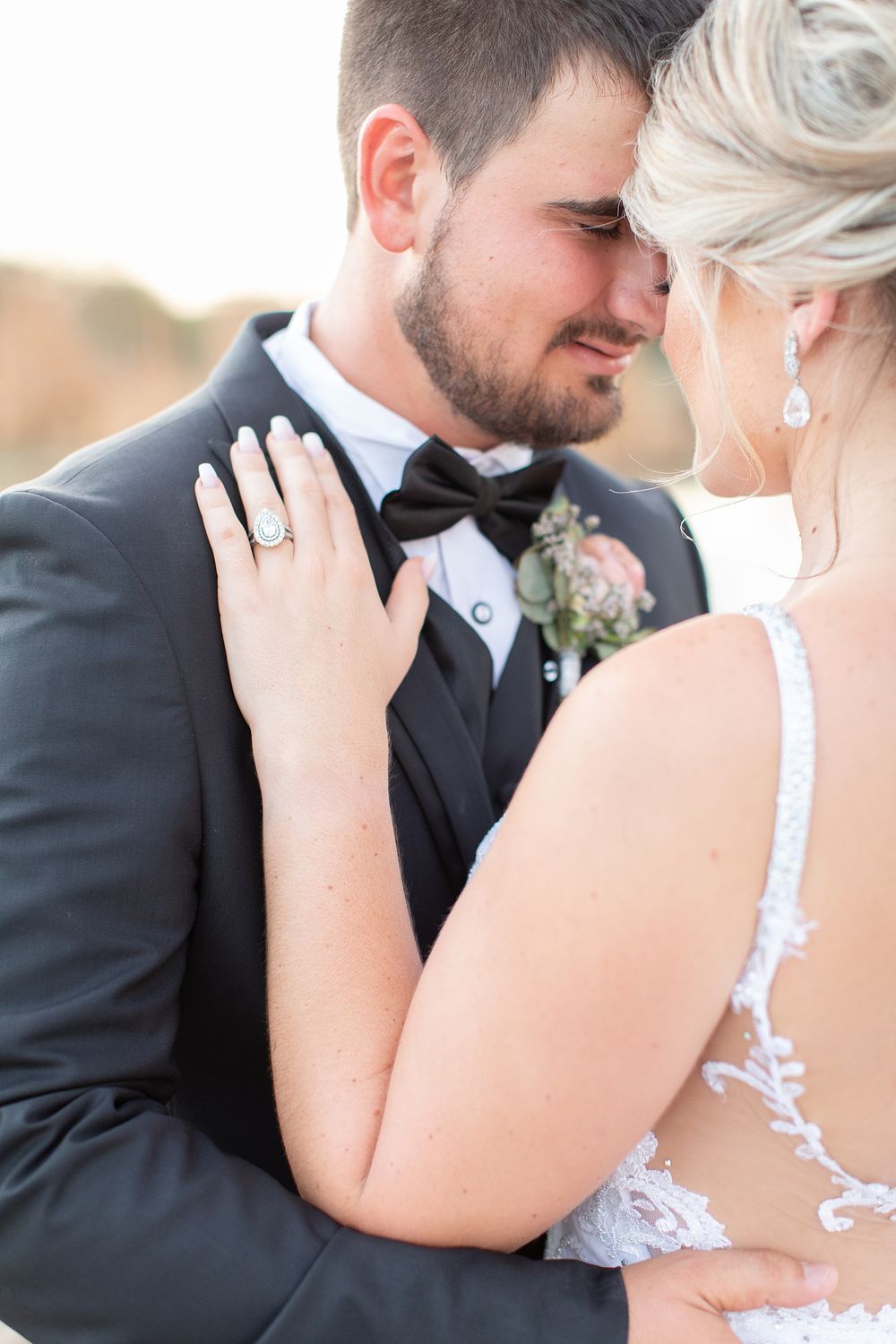 Bride and groom share an intimate moment, bride wearing lace gown and diamond ring, groom in black tuxedo with bow tie.